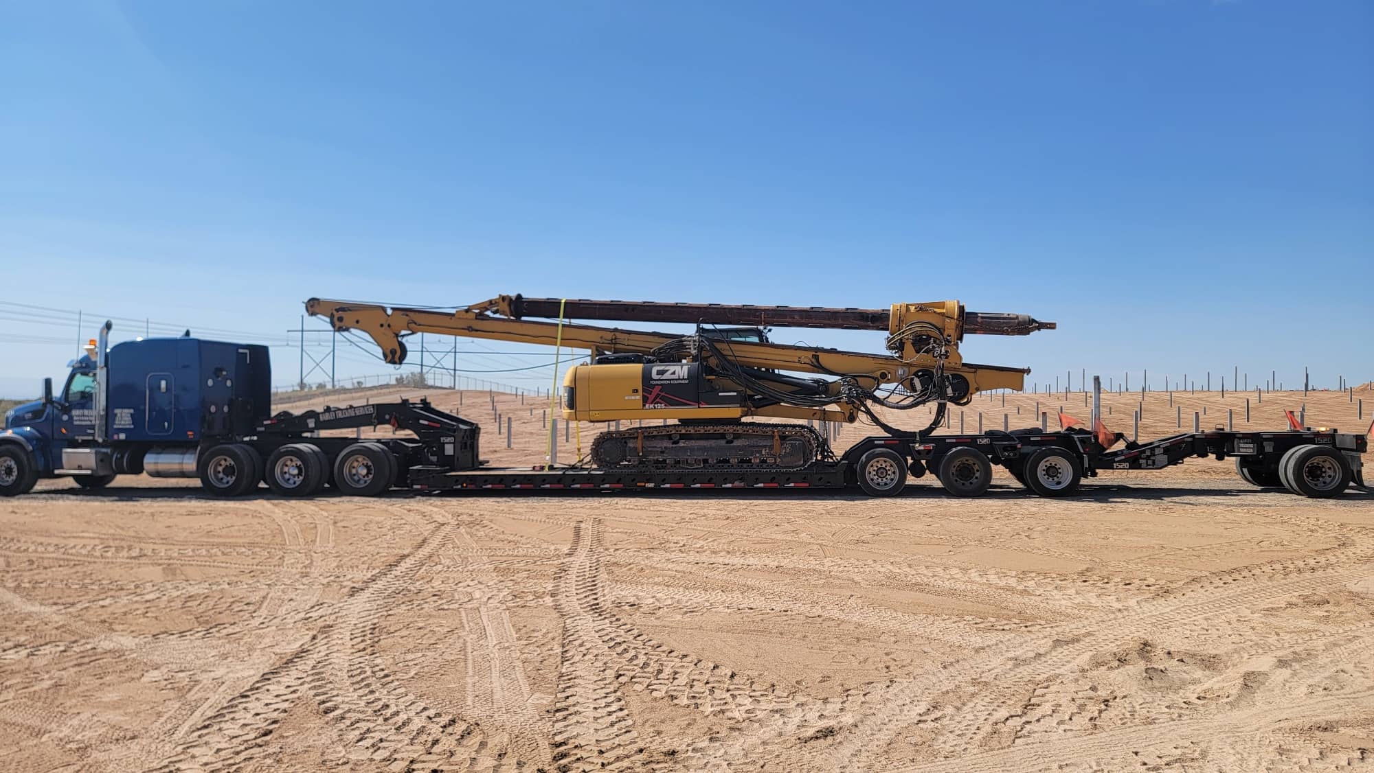 Drill rig secured on a multi-axle RGN trailer in a yard.